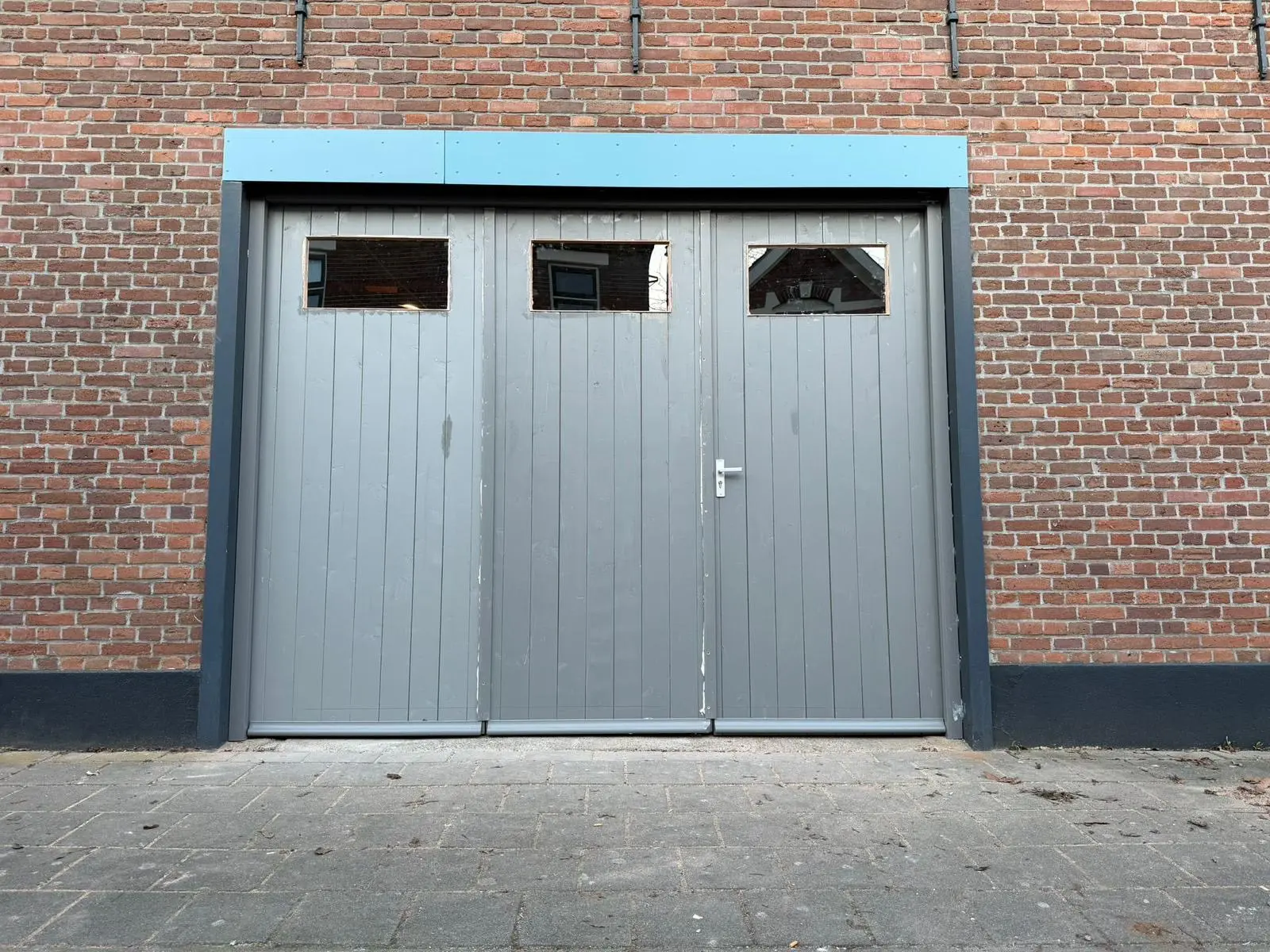 Grijs geschilderde garagedeuren met een lichtblauwe bovenlijst in een bakstenen gevel